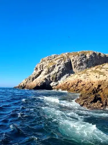 Boat-level view of rugged Marietas Islands cliffs above deep blue Pacific water, with sea spray and seabirds circling the rocky shoreline.