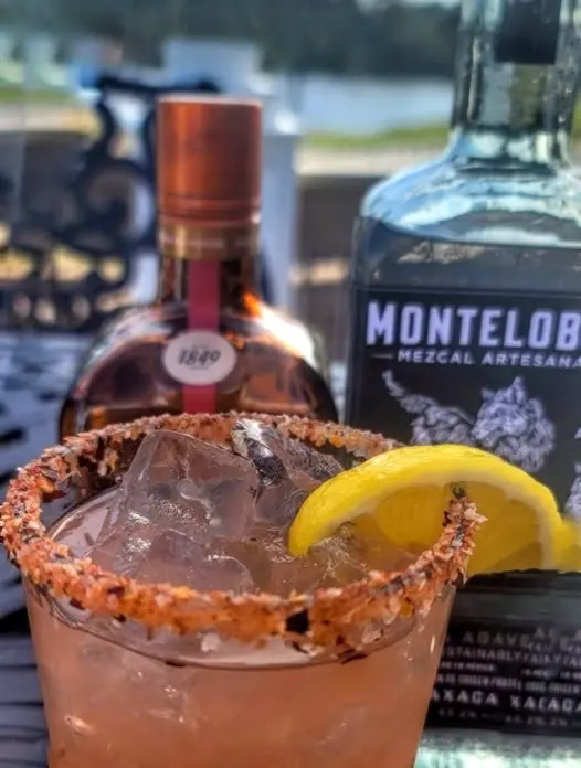 A chili-rimmed mezcal margarita with lemon garnish beside bottles, channeling Riviera Nayarit flavor and golden-hour patio vibes.