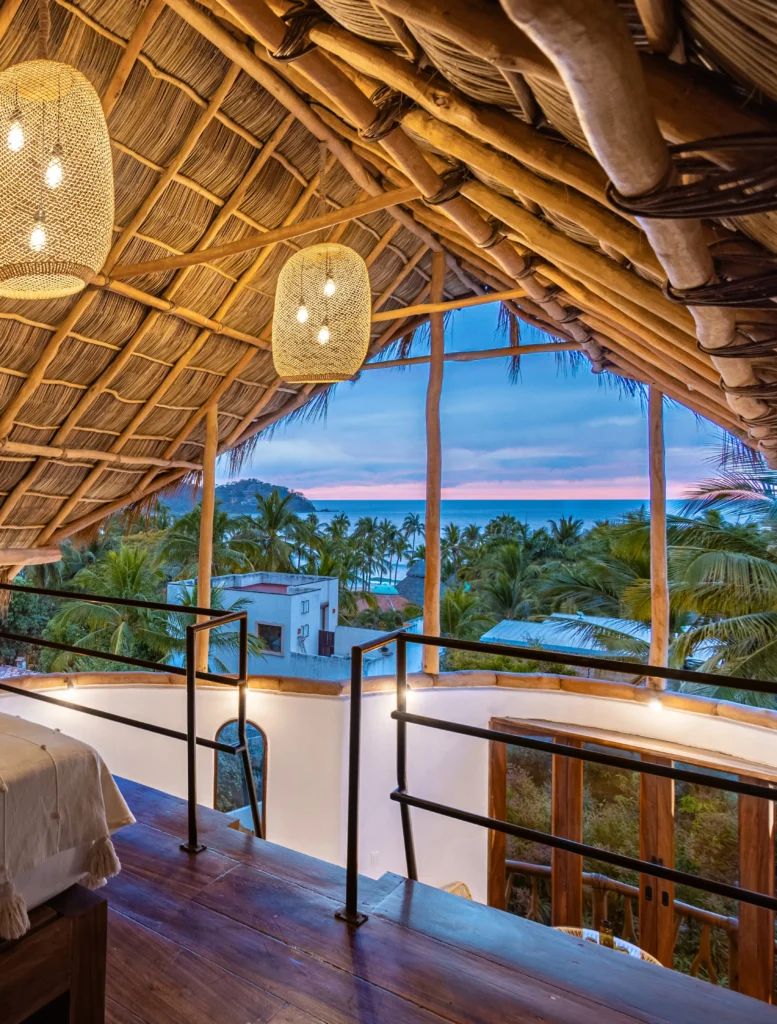 A palapa-roof penthouse loft at Amari Boutique Hotel glows with warm lighting, overlooking palm trees and the ocean along the Costa Banderas.