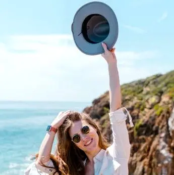 Smiling traveler holding a sun hat overhead on a coastal path with ocean views, dressed for a warm day on the Litibú coast.
