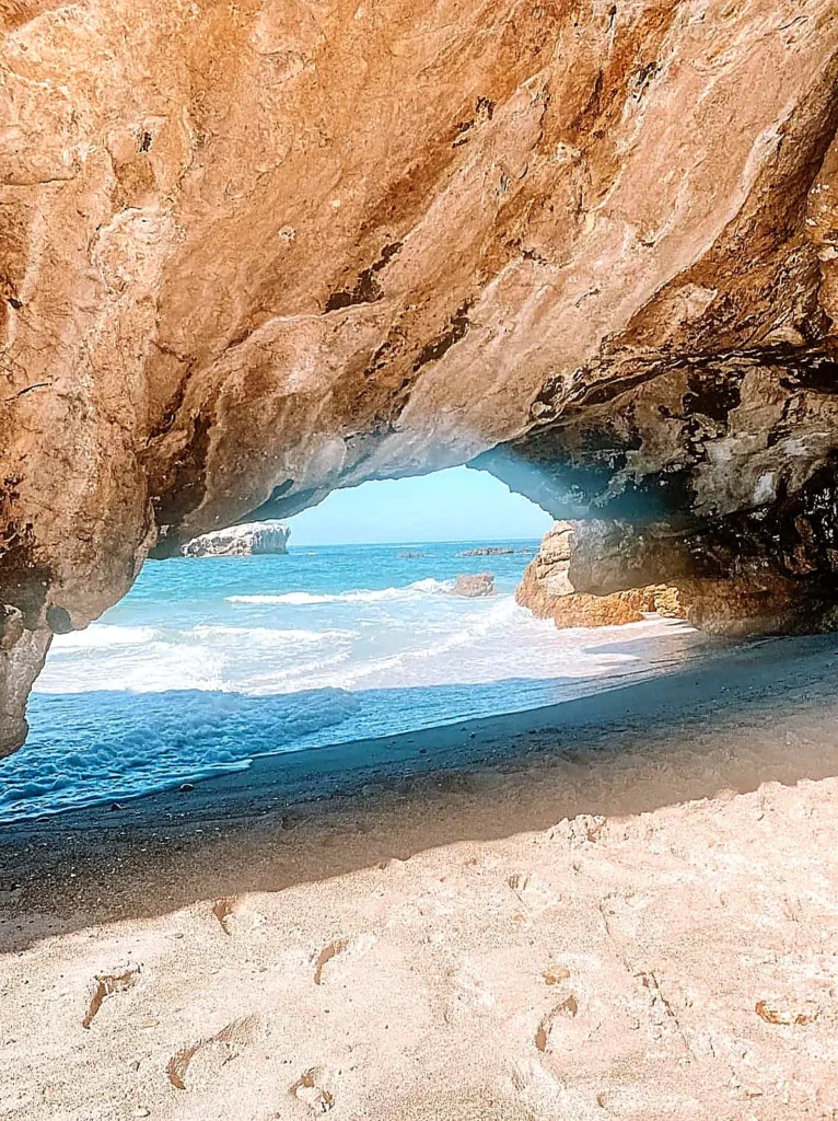 View from beneath a rocky sea cave arch onto a sandy beach with gentle waves and bright blue water at the Marietas Islands on a boating trip from Punta Mita.