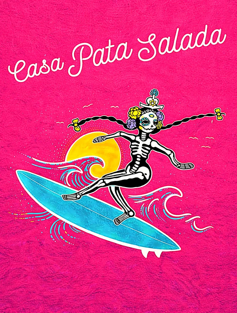 A bright pink mural shows a Día de los Muertos skeleton surfing—street art on Mexico’s Pacific Riviera reading “Casa Pata Salada.”