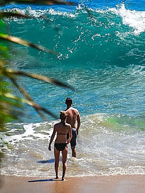 Two people walk toward the shoreline as a turquoise wave rises behind them on Nayarit’s Pacific coast, with wet sand and sea foam.