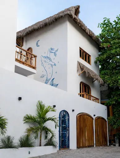 White exterior with palapa-style thatched roof, carved wooden doors, tropical plants, and ocean-inspired mural at Amari Boutique Hotel in Sayulita.
