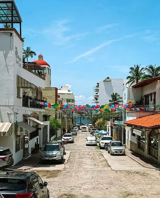 A cobblestone street in Bucerías lined with colorful papel picado, local shops, palm trees, and Pacific Ocean views.