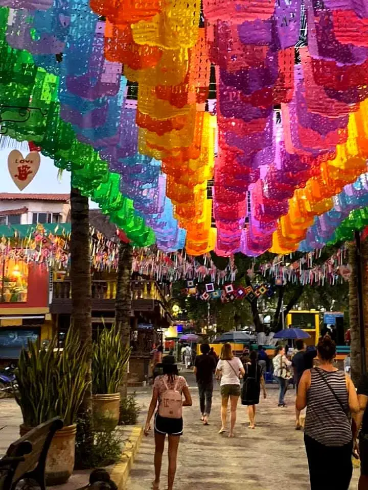 Colorful papel picado banners decorating a lively pedestrian area during an evening celebration.