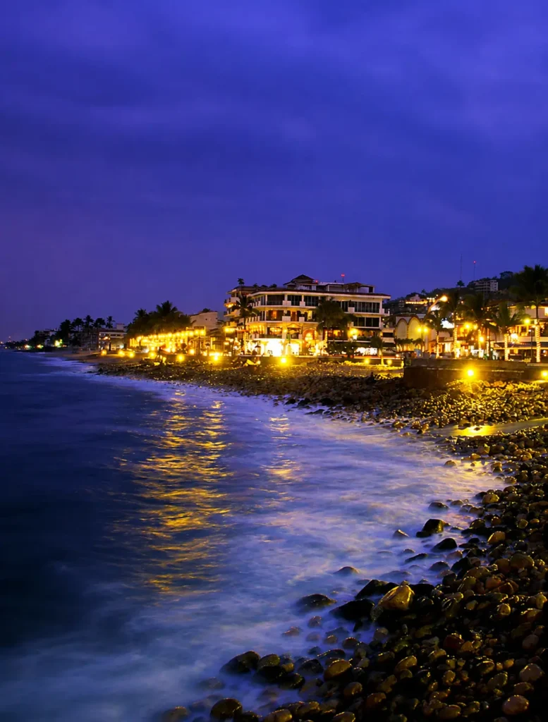 Vista nocturna del malecón de Puerto Vallarta, rodeado de restaurantes, palmeras, edificios iluminados y vistas al océano.