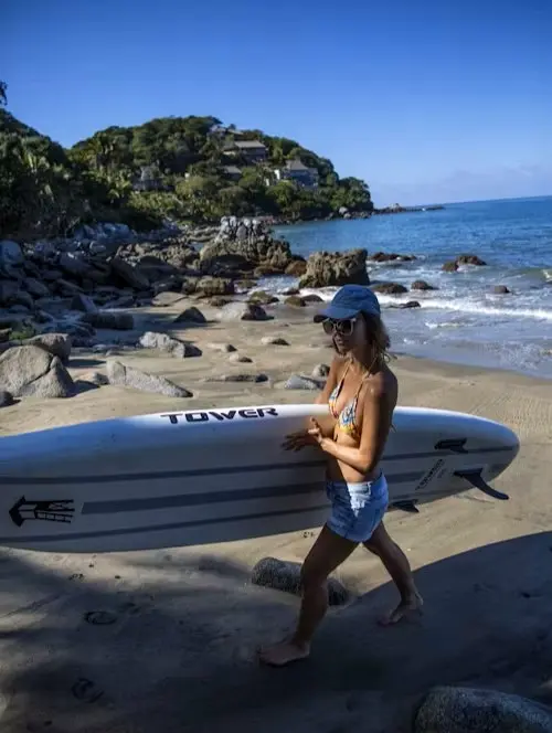 Surfer carrying a board along the South Side shoreline with rocky coastline and hillside homes overlooking the Pacific.
