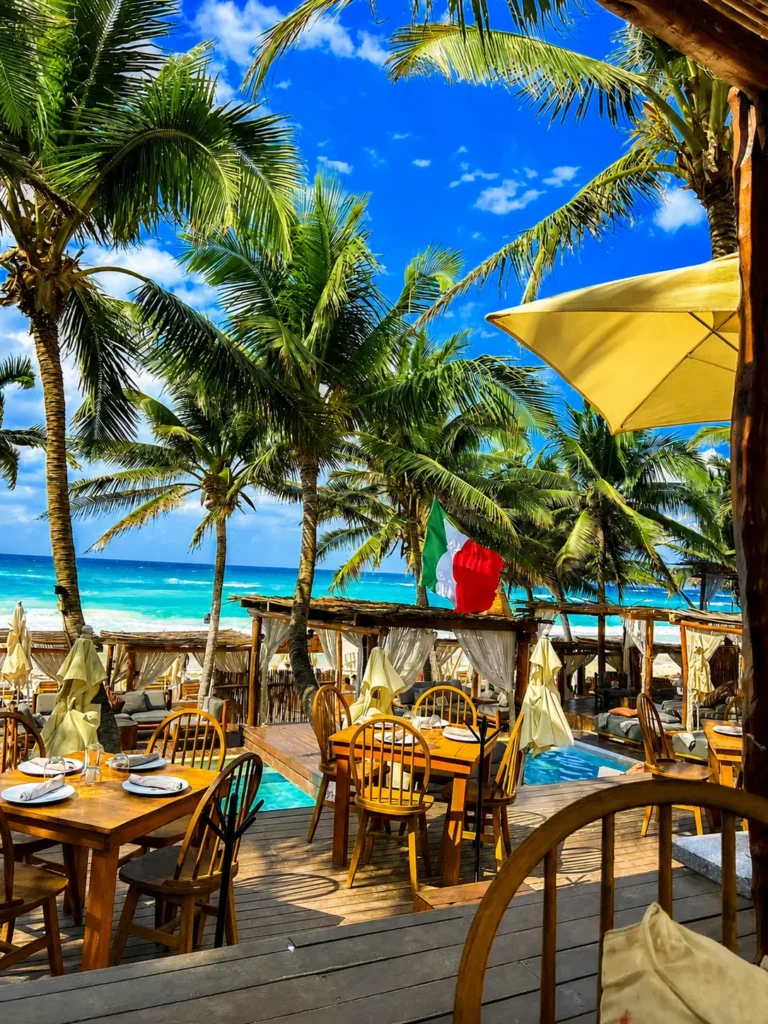 An open-air beachfront restaurant in Tulum overlooking turquoise Caribbean waters, swaying palm trees, and tropical cabanas.